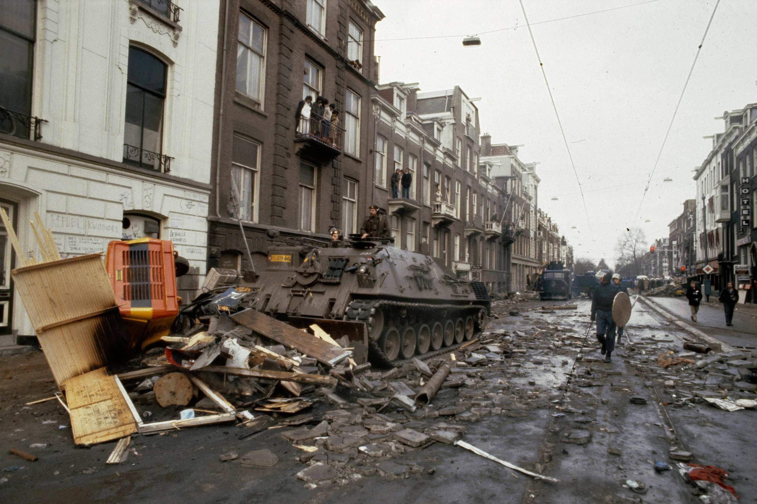 Hout puin rondom een gebouw op de straat wordt opgeruimd door een tank. Er staan mensen op hun balkon toe te kijken en politie is ook aanwezig.