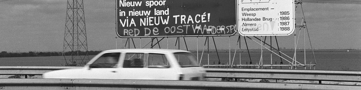 Zwart-wit foto van een auto op de snelweg die langs een bord rijdt waarbij de Flevolijn wordt aangekondigd. Daaronder is met een spuitbus geschreven: Red de Oostvaardsplassen!