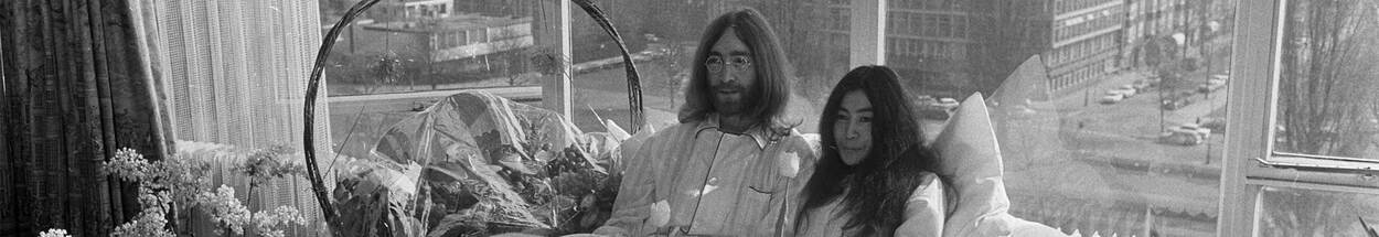 Zwart-wit foto van John Lennon en Yoko Ono in bed. Op de achtergrond is door het raam straten van Amsterdam te zien.