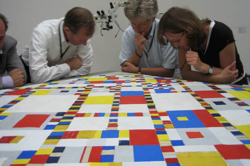 Vier onderzoekers bestuderen de Victory Boogie Woogie van Piet Mondriaan op zaal, met op de voorgrond in close-up het abstracte schilderij.