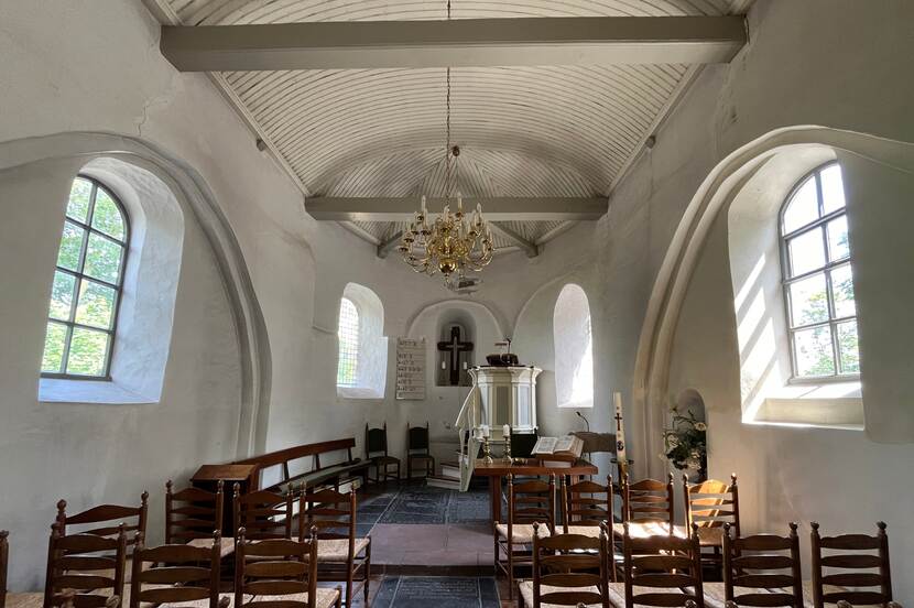 Interieur van een hervormde kerk, gericht richting het altaar. Houten stoelen op de voorgrond, met aan de zijkanten licht dat door de ramen schijnt. In het witte gesteente van de muren zijn scheuren zichtbaar.