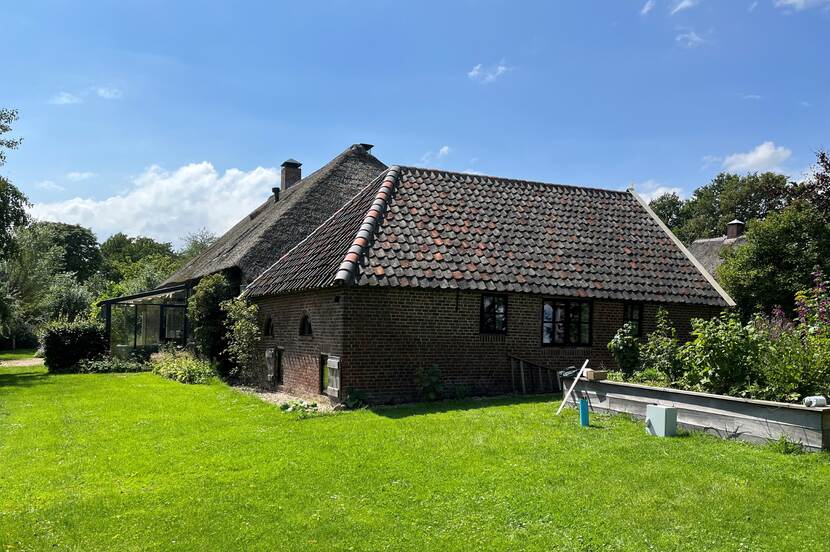 Foto met hoekaanzicht van de rijksmonumentale boerderij in Rossum. Op de voorgrond een grasveld, om de boerderij heen is begroeiing te zien.