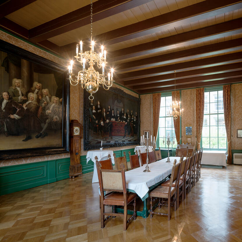 Collegezaal met een lange tafel in het midden, een kroonluchter erboven en een aantal schilderijen aan de muur