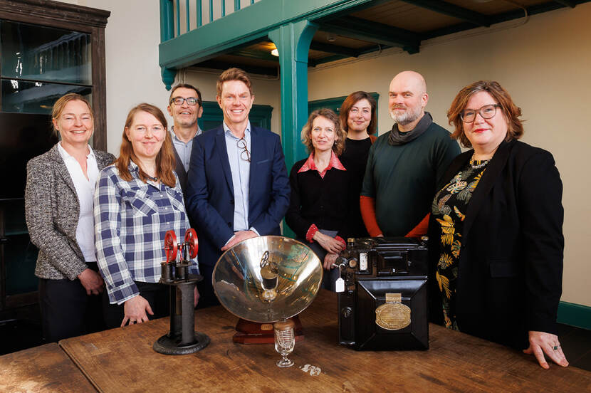 Acht mensen poseren achter een tafel waarop voorwerpen uit wetenschappelijke collecties zijn uitgestald.