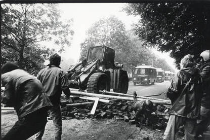 Een zwart-wit afbeelding van een tractor die een barricade opruimt met daar omheen nog een paar mannen.